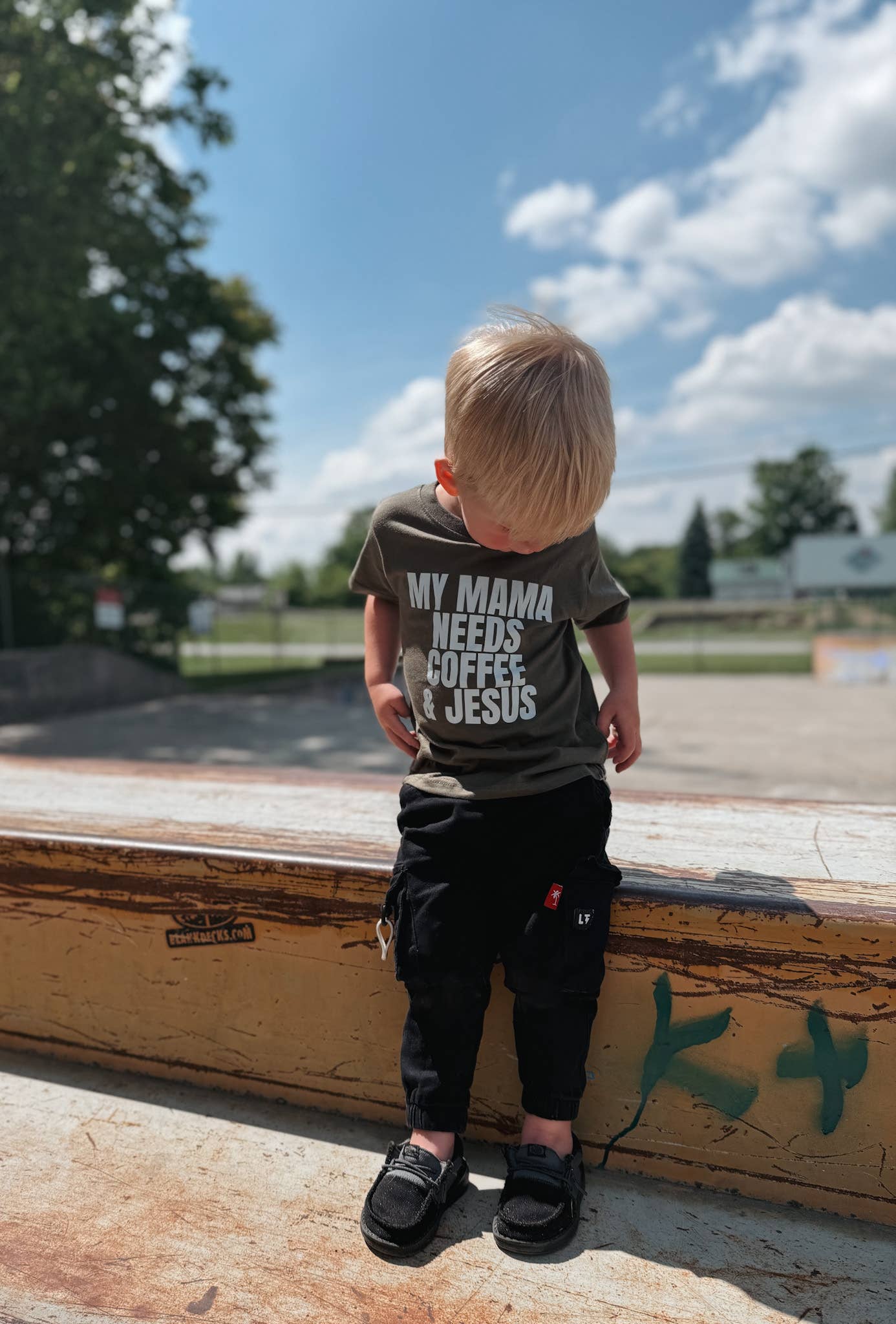 Child wearing a shirt with text, standing on a ledge outdoors with trees and sky in the background.