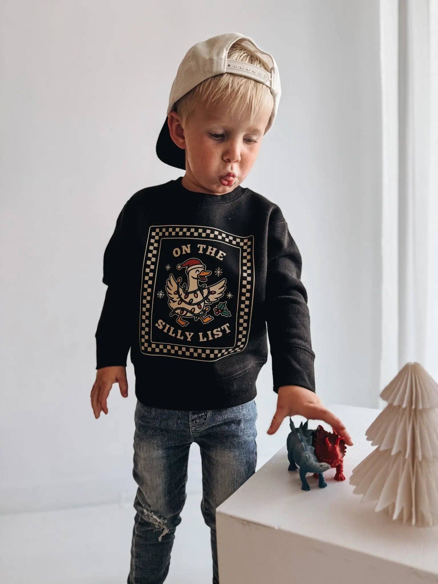 Child wearing a black sweatshirt with graphic design and text, standing next to small figurines on a white surface.