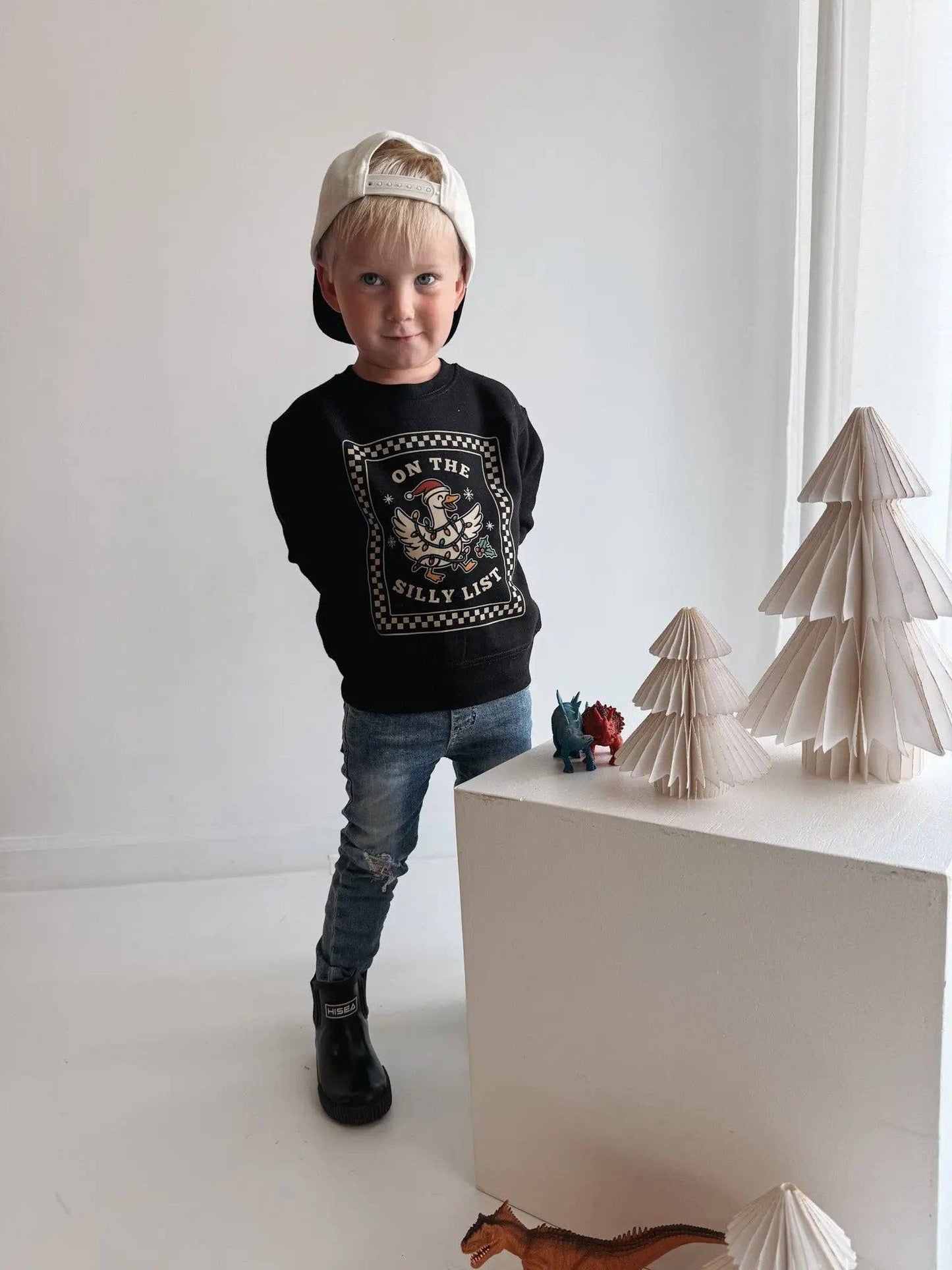 Child wearing a black sweater with text and graphics, standing next to decorative paper trees on a white surface.