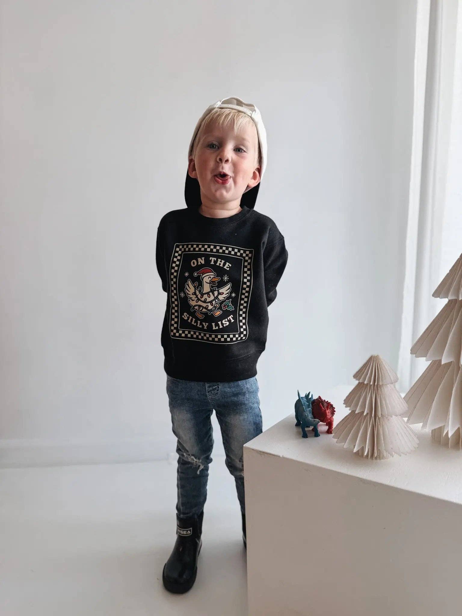 Child wearing a black sweatshirt with text and graphics, standing in a minimalistic room with a white wall and a small table with decorative items.