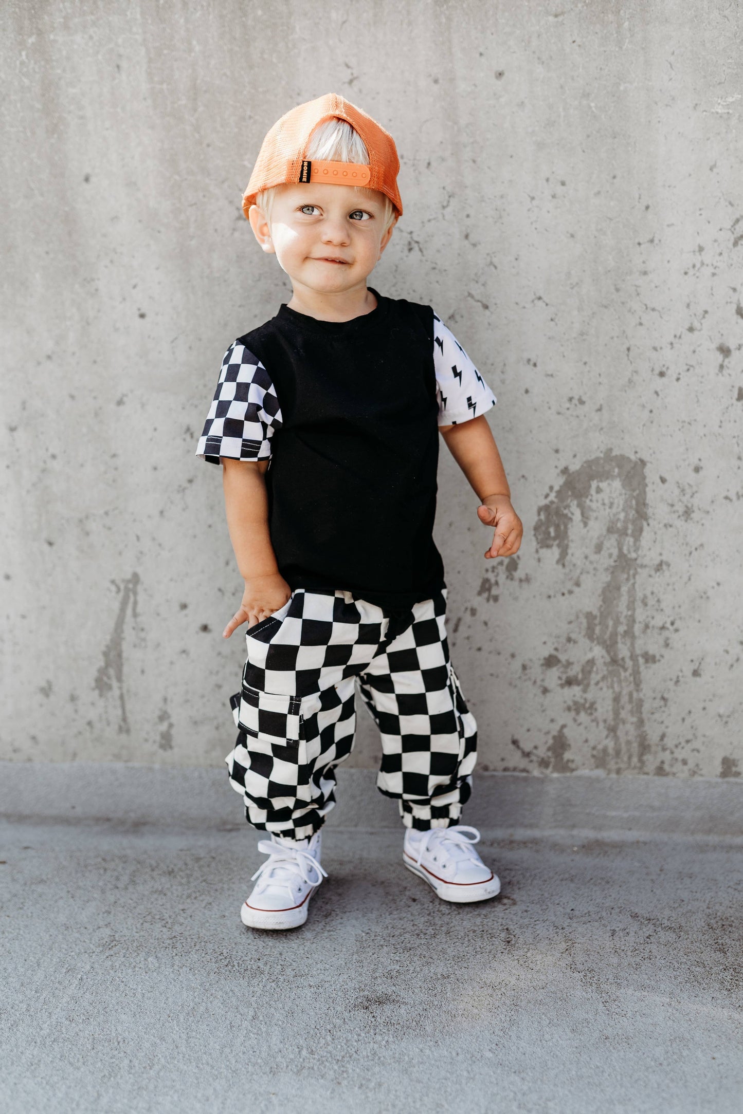 Child wearing a black and white checkered outfit with an orange cap against a concrete wall.