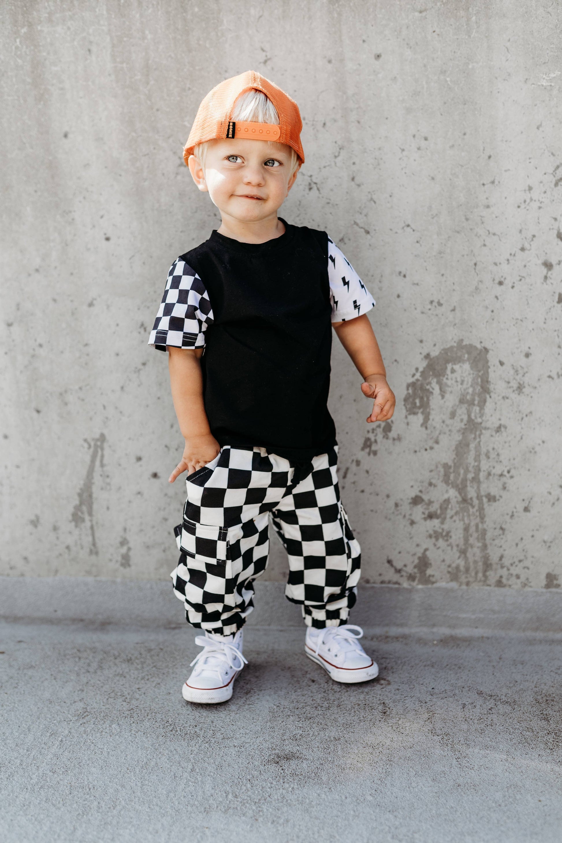 Child wearing a black and white checkered outfit with an orange cap against a concrete wall.