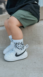 Child's legs wearing white sneakers with black swoosh logo and checkered socks with a smiley face design.
