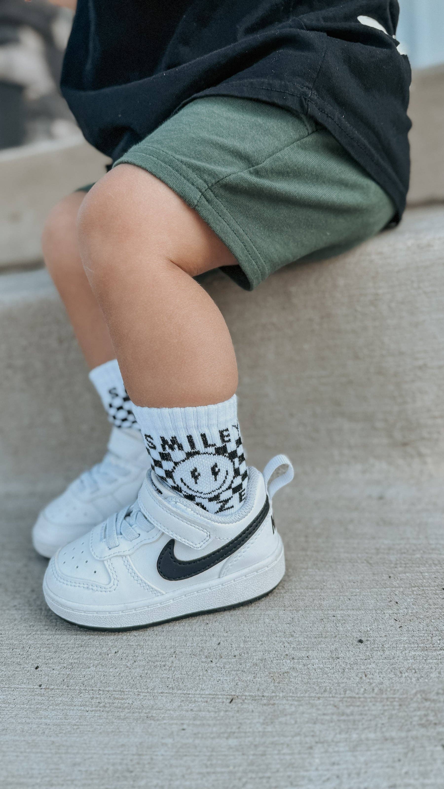 Child's legs wearing white sneakers with black swoosh logo and checkered socks with a smiley face design.