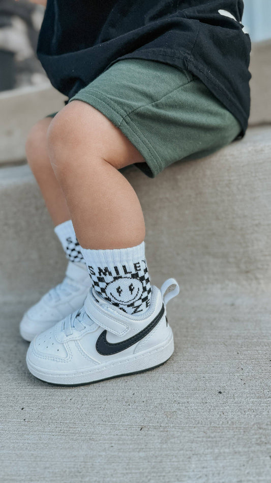 Child's legs wearing white sneakers with black swoosh logo and checkered socks with a smiley face design.