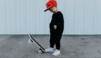 Child wearing a red cap and black outfit standing next to a skateboard on a concrete surface.