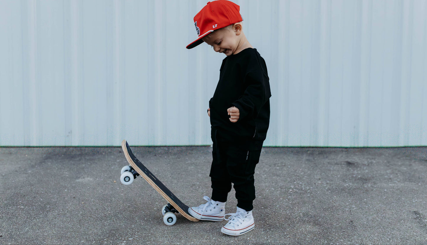 Child wearing a red cap and black outfit standing next to a skateboard on a concrete surface.