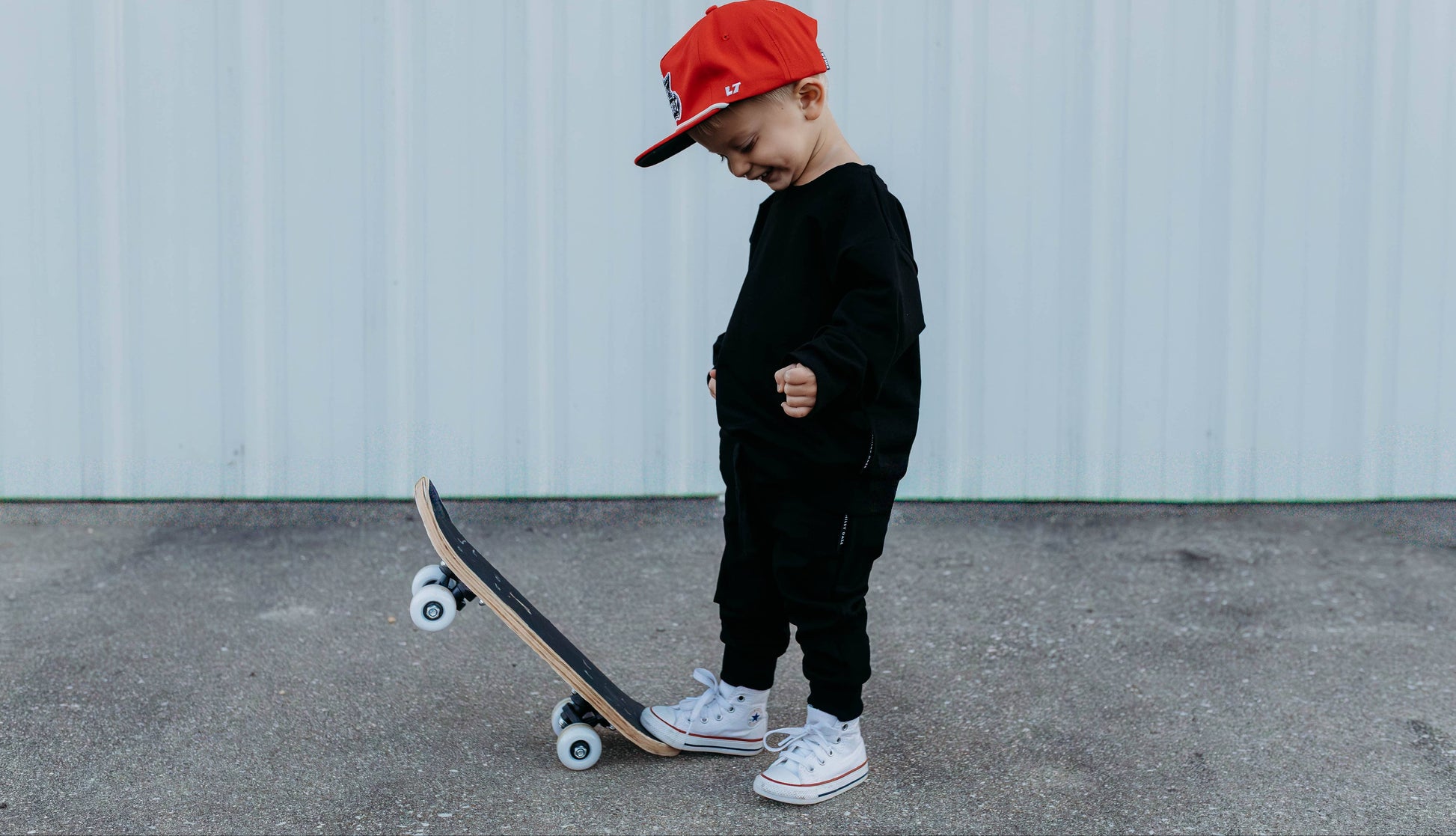 Child wearing a red cap and black outfit standing next to a skateboard on a concrete surface.