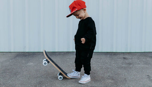 Child wearing a red cap and black outfit standing next to a skateboard on a concrete surface.