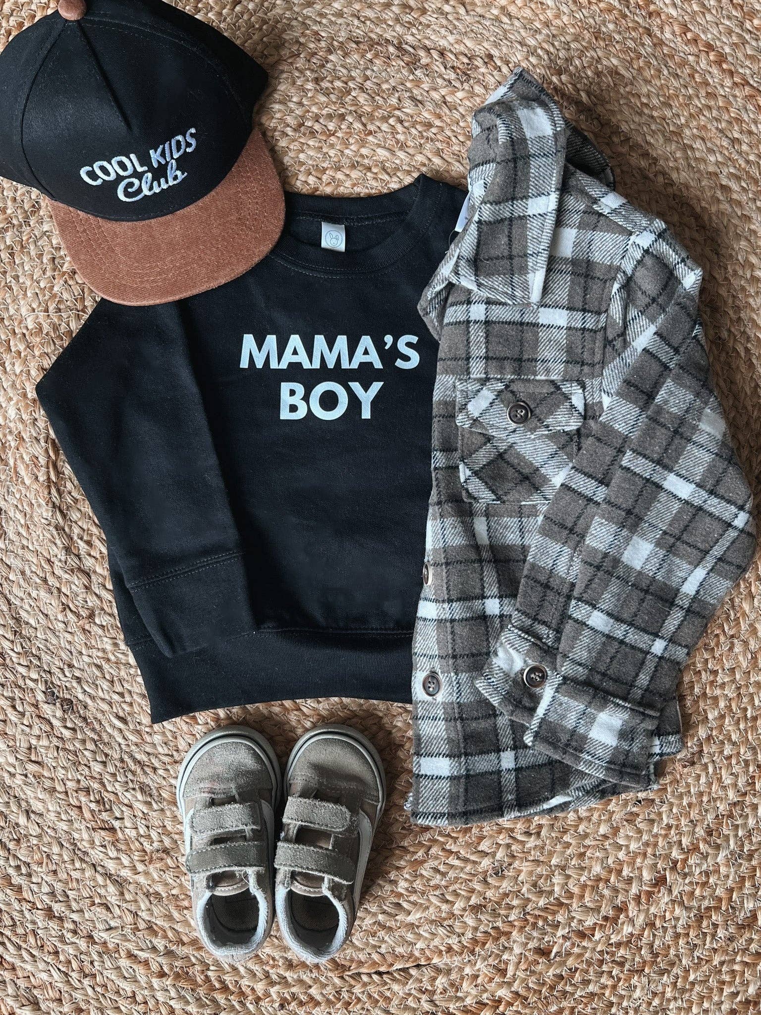 Black sweatshirt with 'MAMA'S BOY', plaid shirt, cap, and shoes on a textured surface