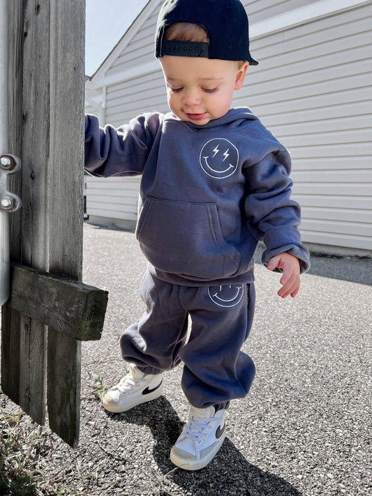 Child wearing a blue hoodie and pants with a smiley face design, standing next to a wooden post.