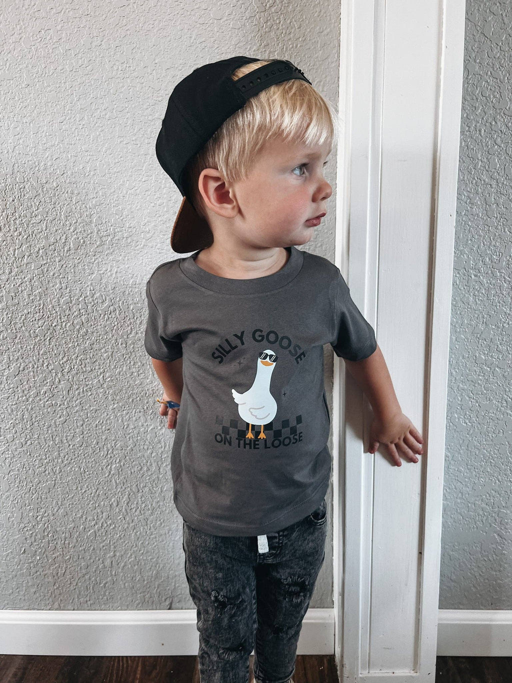 Child wearing a gray t-shirt with a graphic design and text, standing against a plain wall.
