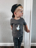 Child wearing a gray t-shirt with a graphic design and text, standing against a plain wall.