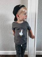 Child wearing a gray t-shirt with a graphic design and text, standing against a plain wall.