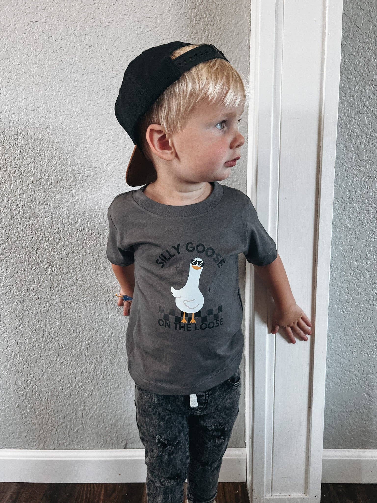 Child wearing a gray t-shirt with a graphic design and text, standing against a plain wall.